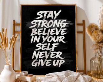 Puede incluir: Un póster enmarcado con fondo negro y texto blanco: "Stay Strong, Believe in Yourself, Never Give Up". El póster se exhibe sobre un caballete de madera. Elementos decorativos, como un jarrón y plantas secas, están en el fondo.