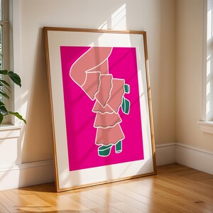 Könnte beinhalten: Ein gerahmter Kunstdruck mit einer stilisierten Illustration einer Figur in einem rosa Kleid mit grünen Schuhen vor einem leuchtend pinkfarbenen Hintergrund. Das Kunstwerk wird in einem Raum mit natürlichem Licht und Holzboden ausgestellt.
