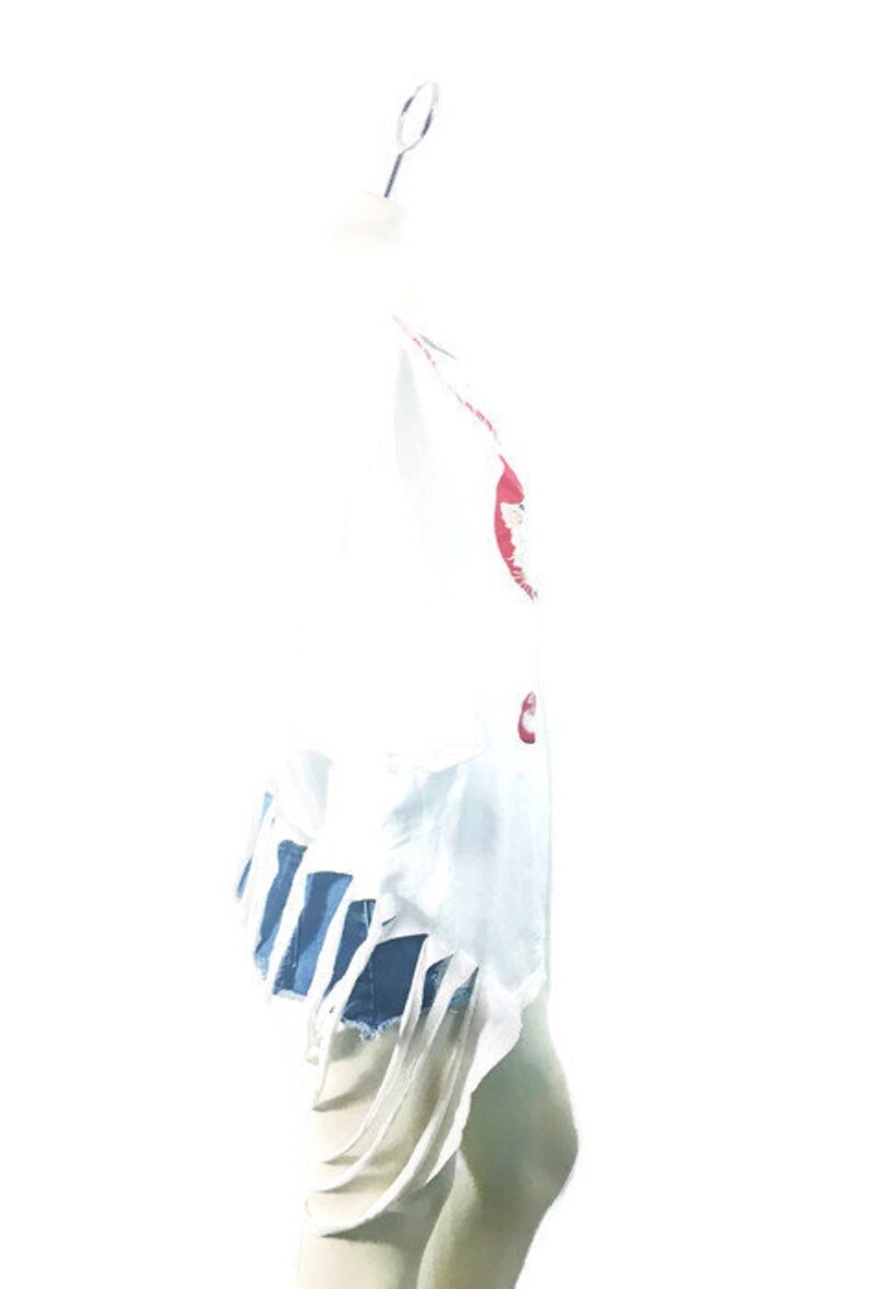 Puede incluir: Camiseta blanca con bordes deshilachados y un estampado gr&aacute;fico rojo y azul. La camiseta est&aacute; en un maniqu&iacute; blanco.