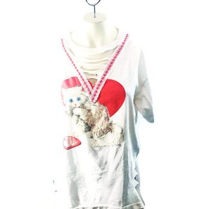 Puede incluir: Camiseta blanca con un escote desgastado y un coraz&oacute;n rojo con un gr&aacute;fico de gatito. El coraz&oacute;n tiene un borde de encaje rosa y blanco.