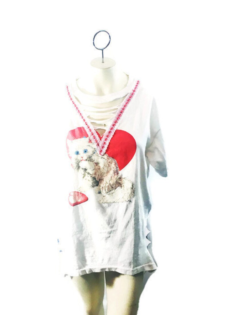Puede incluir: Camiseta blanca con un escote desgastado y un coraz&oacute;n rojo con un gr&aacute;fico de gatito. El coraz&oacute;n tiene un borde de encaje rosa y blanco.