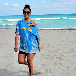 Könnte beinhalten: Eine Frau mit Sonnenbrille und einem blauen Batik-T-Shirt mit dem Text "Here I come to save the day" am Strand.