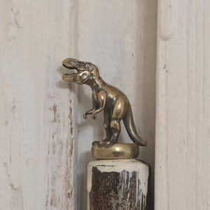 Könnte beinhalten: Kleine, messingfarbene Dinosaurierfigur auf einem zylindrischen Sockel mit weiß-braunem Used-Finish. Der Dinosaurier ist mit offenem Maul dargestellt, auf einer dunklen, möglicherweise hölzernen Oberfläche.