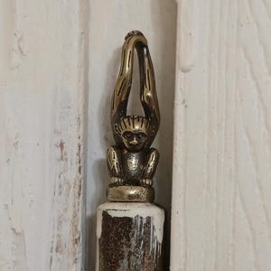May include: A decorative brass hinge featuring a small monkey figure with its arms raised. The hinge is mounted on a white wooden door, adding a unique and artistic touch to the door's hardware. The monkey figure is detailed and adds a whimsical element.