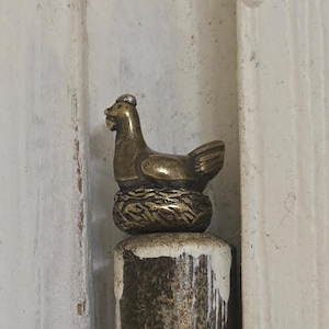 May include: A bronze-coloured hen figurine on a nest, mounted on a cylindrical post. The post has a textured, aged appearance with a white and brown finish. The background is a weathered, light-coloured wooden surface.