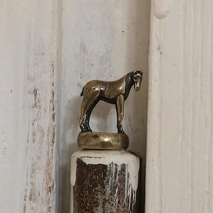 Puede incluir: Una pequeña figura de caballo de color bronce se encuentra sobre un objeto cilíndrico y texturizado. El caballo tiene patas detalladas y una melena definida. La base de la figura es un disco circular dorado. El fondo es una superficie blanca texturizada.