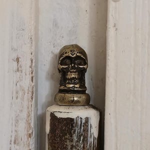 May include: A decorative bottle stopper with a skull design. The skull is bronze-colored and sits atop a white and bronze-colored stopper. The background is a white, textured surface.