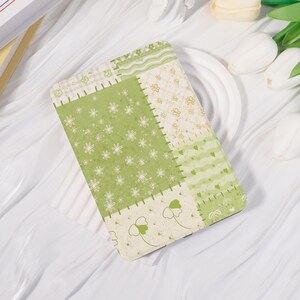 Könnte beinhalten: Eine rechteckige Tablethülle mit einem Patchwork-Design in Grün-, Creme- und Weißtönen. Das Muster besteht aus Abschnitten mit weißen Blumenprints, kleinen Schmetterlingen, wellenförmigen Linien und Herzmotiven, die einen visuellen Effekt erzeugen.