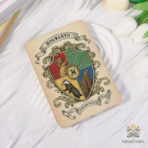 Puede incluir: Una funda rectangular beige con el escudo de Hogwarts. El escudo está dividido en cuatro secciones, cada una con un animal y un color diferentes. La palabra "HOGWARTS" está impresa en la parte superior en una pancarta.