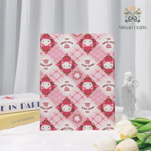 May include: A pink and white Hello Kitty notebook with a repeating pattern of Hello Kitty faces, roses, and the words "Hello Kitty". The notebook is standing upright, with books and a small statue in the background.