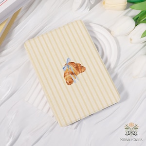 Op de afbeelding: Een rechthoekige hoes met een crème- en beige gestreept patroon. Een aquarel illustratie van een croissant met een blauw lint is gecentreerd op de hoes. De hoes is waarschijnlijk voor een e-reader of tablet.