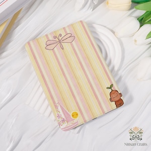 May include: A rectangular notebook with a striped pattern in yellow and pink. The notebook features a pink dragonfly, a cartoon sailboat, a sun, and a cartoon character. The notebook is a pastel color.