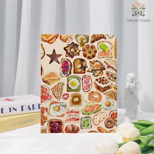 May include: A notebook with a beige background featuring a collage of watercolor illustrations of various types of toast, sandwiches, and pastries. A brown star is also included in the design. The notebook is a rectangular shape.