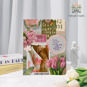 Op de afbeelding: Een decoratief notitieboek met een collage-ontwerp met roze bloemen, een slapende kat en de tekst "it's okay to not be okay". Het notitieboek bevat ook een bioscoopkaartje en de zin "you're precious I hope you know that."