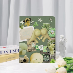 May include: A sage green notebook cover featuring a collage of whimsical illustrations including two beige figures forming a heart shape, green frogs, a digital pet, a camera, and the word LOVE in block letters, accented with white stars and doodles.