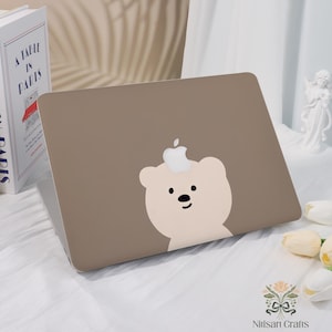 May include: A taupe laptop case featuring a cartoon bear design with a white apple on its head. The bear has a light beige face with black eyes and a black nose. The laptop case is on a white surface.