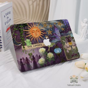 May include: A laptop case with a tarot card design. The case has a purple background with a sun, stars, moon, and fairy illustrations. The text "THE LOVERS" is displayed on a banner. White roses and a green frog plushie are also visible.