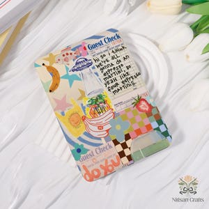 May include: A colorful phone case with a collage design featuring a banana, an alien spaceship, and text that reads "Guest Check." The case has a playful aesthetic with a mix of patterns and illustrations, including a strawberry and flowers.