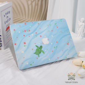 May include: A light blue laptop case with a watercolour design featuring a green sea turtle, white Apple logo, and scattered pink and white flowers. The case has a wavy pattern and is on a white surface.