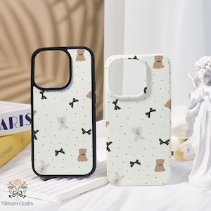 May include: Two phone cases with a white background, featuring a repeating pattern of brown teddy bears, black bows, and small black dots. One case has a black border, the other has a white border. The cases are for a smartphone.