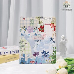 Op de afbeelding: Een decoratief notitieboek met een collage-ontwerp. De omslag heeft een mix van patronen, waaronder ruitjes en bloemmotieven, met de tekst "Slow down, you're doing fine" en "and finally she chose herself." Het ontwerp bevat vlinders, aardbeien en een stadsgezicht.