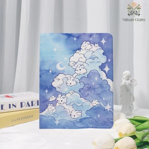 Op de afbeelding: Een notitieboek met een aquarel illustratie van een blauwe lucht vol zachte wolken, sterren en halve manen. Witte cartoon konijntjes nestelen in de wolken. Het notitieboek wordt getoond met boeken en een klein beeldje.