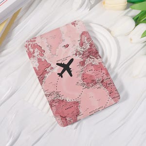 May include: A rectangular e-reader case featuring a pink world map print with a black airplane silhouette flying over the North Atlantic Ocean. The design includes dashed flight paths and text labels for the North Atlantic and South Atlantic Oceans.