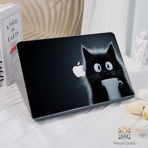 Könnte beinhalten: Eine schwarze Laptop-Hülle mit einer Cartoon-Katze, die eine weiße Tasse hält. Das Apple-Logo ist sichtbar. Der Laptop liegt auf einer weißen Oberfläche. Das Design ist verspielt.