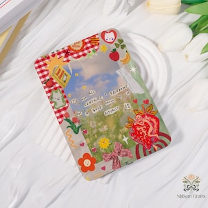 May include: A rectangular, decorative resin piece with a whimsical design. It features a sky and flower scene, surrounded by a red and white checkered border. Adorned with stickers of fruit, cats, and the text "It's not all sunshine & rainbows but a good amount of it actually is."