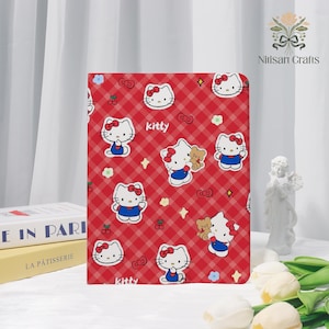 Puede incluir: Cuaderno a cuadros rojo y blanco con múltiples ilustraciones de Hello Kitty. El cuaderno tiene un fondo rojo con líneas blancas y la palabra "Kitty" está impresa en la portada. Un artículo lindo y colorido.