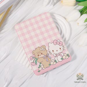 Peut inclure: Un étui rose et blanc à motif vichy avec un ours de dessin animé et Hello Kitty. L'étui a une bordure rose et est décoré de fleurs. L'étui est de forme rectangulaire.