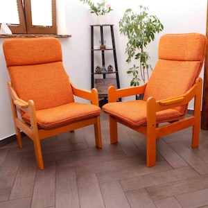 May include: Two mid-century modern armchairs with orange upholstery and light wood frames. The chairs have high backs and armrests, and are placed on a dark wood floor. The chairs are in a well-lit room.