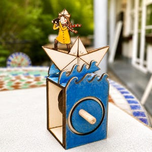 May include: A wooden mechanical toy featuring a figure in a yellow coat and striped scarf standing on a paper-style boat. The boat sits atop blue wave cutouts on a rectangular base with a circular side crank handle for manual operation.