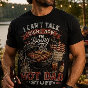 May include: Black t-shirt with a barbecue-themed graphic. The design includes a grill with steaks, an American flag, and the text "I CAN'T TALK RIGHT NOW I'M Doing HOT DAD STUFF". The shirt also features a bottle of BBQ sauce and a glass of iced tea.