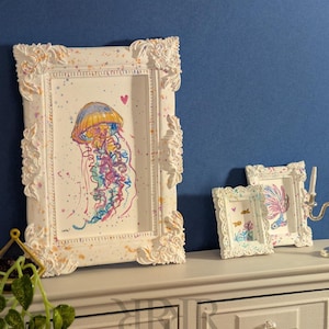 May include: White framed art prints featuring watercolor jellyfish and fish illustrations. The frames have ornate detailing and a distressed finish with colorful paint splatters. The art is displayed on a gray dresser against a blue wall.