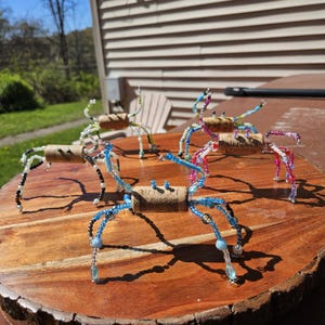 May include: A collection of handmade beaded crab sculptures. Each crab features a cork body and wire legs adorned with colorful beads in shades of blue, green, black, and pink. The crabs are displayed on a wooden surface.