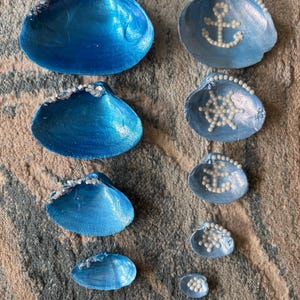 Puede incluir: Una colección de conchas marinas pintadas de varios tamaños. Las conchas están pintadas en tonos azules y adornadas con adornos blancos. Algunas conchas presentan un ancla o un diseño de rueda hecho de pequeñas cuentas blancas, creando un tema náutico.
