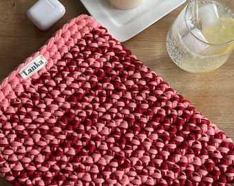 May include: A handmade, two-tone crochet laptop sleeve in shades of pink and burgundy. The sleeve features the brand name "Lanka" on a small label. The sleeve is shown on a wooden surface, with a lit candle, a glass of water, and a pair of white wireless earbuds.