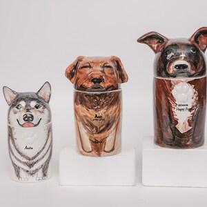 May include: Three ceramic dog urns, each with a unique painted design. The urns feature a husky, a golden retriever, and a brown dog. The husky urn has the name "Andie". The golden retriever urn has the name "Ace". The brown dog urn has the text "Winnie Happy Forever".