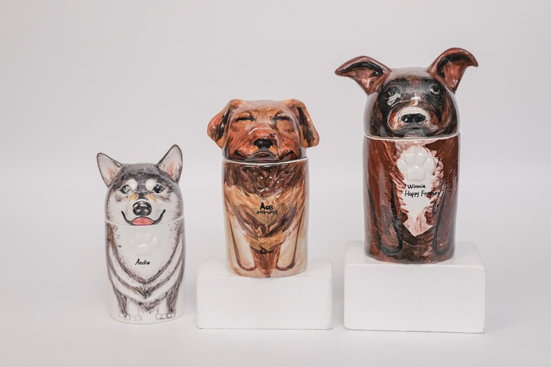 May include: Three ceramic dog urns, each with a unique painted design. The urns feature a husky, a golden retriever, and a brown dog. The husky urn has the name "Andie". The golden retriever urn has the name "Ace". The brown dog urn has the text "Winnie Happy Forever".