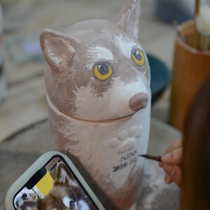May include: A ceramic dog-shaped urn with a lid, painted in shades of brown and white, with yellow eyes. The urn has paw prints and the name "NINA 2016 20" written on it. A phone displays a photo of a dog.