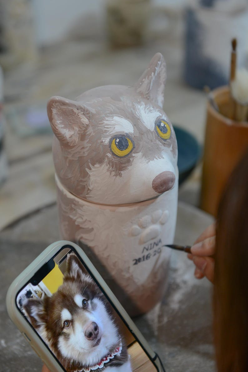 May include: A ceramic dog-shaped urn with a lid, painted in shades of brown and white, with yellow eyes. The urn has paw prints and the name "NINA 2016 20" written on it. A phone displays a photo of a dog.