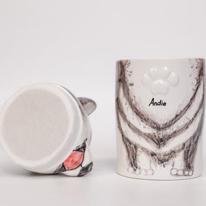 May include: A white ceramic cat-themed mug and a matching cat-shaped object. The mug features a cat's rear end design with the name "Andie" and a paw print. The other object is a cat head with a red and black design.