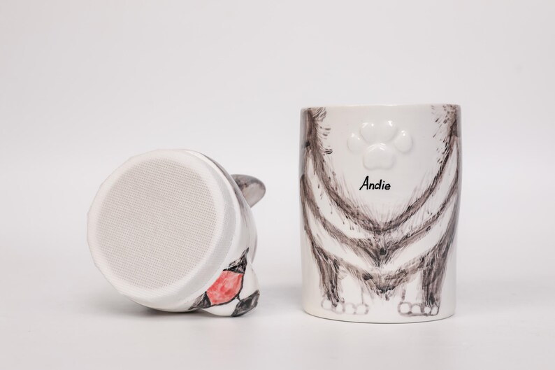 May include: A white ceramic cat-themed mug and a matching cat-shaped object. The mug features a cat's rear end design with the name "Andie" and a paw print. The other object is a cat head with a red and black design.