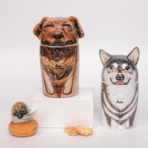 May include: Two ceramic dog-shaped containers, one brown and one black and white, with lids shaped like dog heads. The brown dog container has the name "Ace" on it. The black and white dog container has the name "Andie" on it. A small pastry is in the foreground.