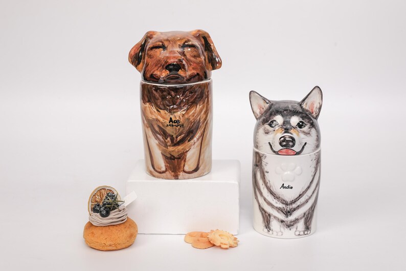 May include: Two ceramic dog-shaped containers, one brown and one black and white, with lids shaped like dog heads. The brown dog container has the name "Ace" on it. The black and white dog container has the name "Andie" on it. A small pastry is in the foreground.