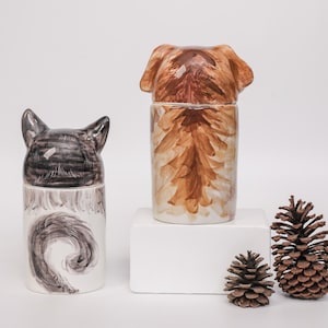 May include: Two ceramic jars, one with a cat head and tail design, the other with a dog head and body design. Two pine cones are next to the jars. The jars are white with brown and black painted details.