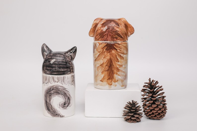 May include: Two ceramic jars, one with a cat head and tail design, the other with a dog head and body design. Two pine cones are next to the jars. The jars are white with brown and black painted details.