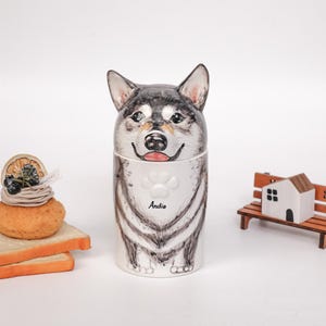 May include: A ceramic dog-shaped container with a black and gray design, featuring the name "Andie" on a white bib. Beside it are miniature wooden houses and a bench, along with a small dessert on toast.