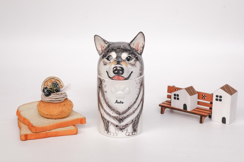 May include: A ceramic dog-shaped container with a black and gray design, featuring the name "Andie" on a white bib. Beside it are miniature wooden houses and a bench, along with a small dessert on toast.